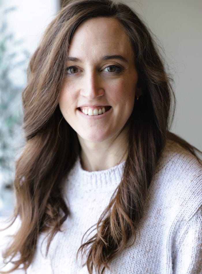 Erin Thomas smiling, in a cream knit sweater by a window